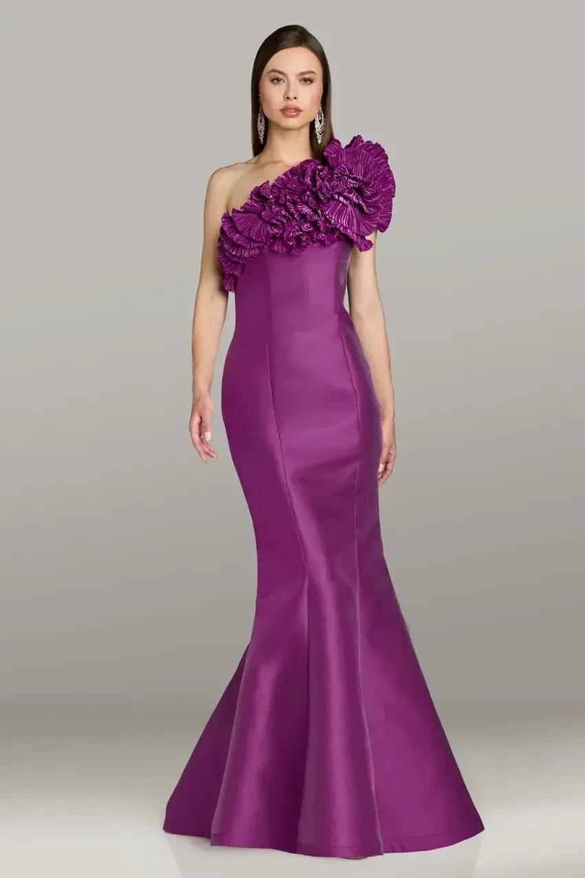 A model wearing a sleek, one-shoulder purple evening gown with a dramatic floral embellishment on the shoulder. The gown has a fitted silhouette that flares out at the bottom.