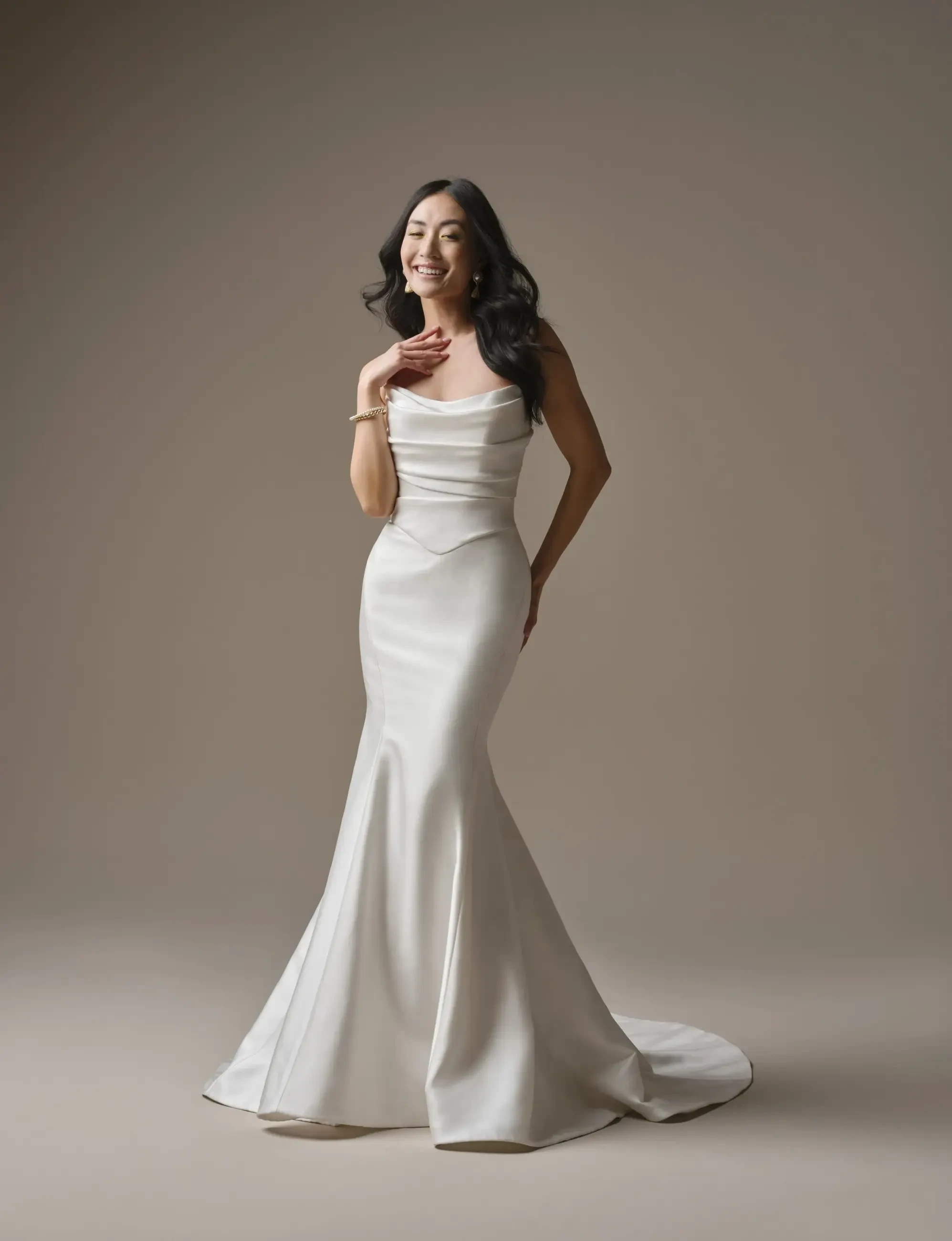A woman in an elegant, fitted white gown with a mermaid silhouette, smiling and posing gracefully against a neutral background.
