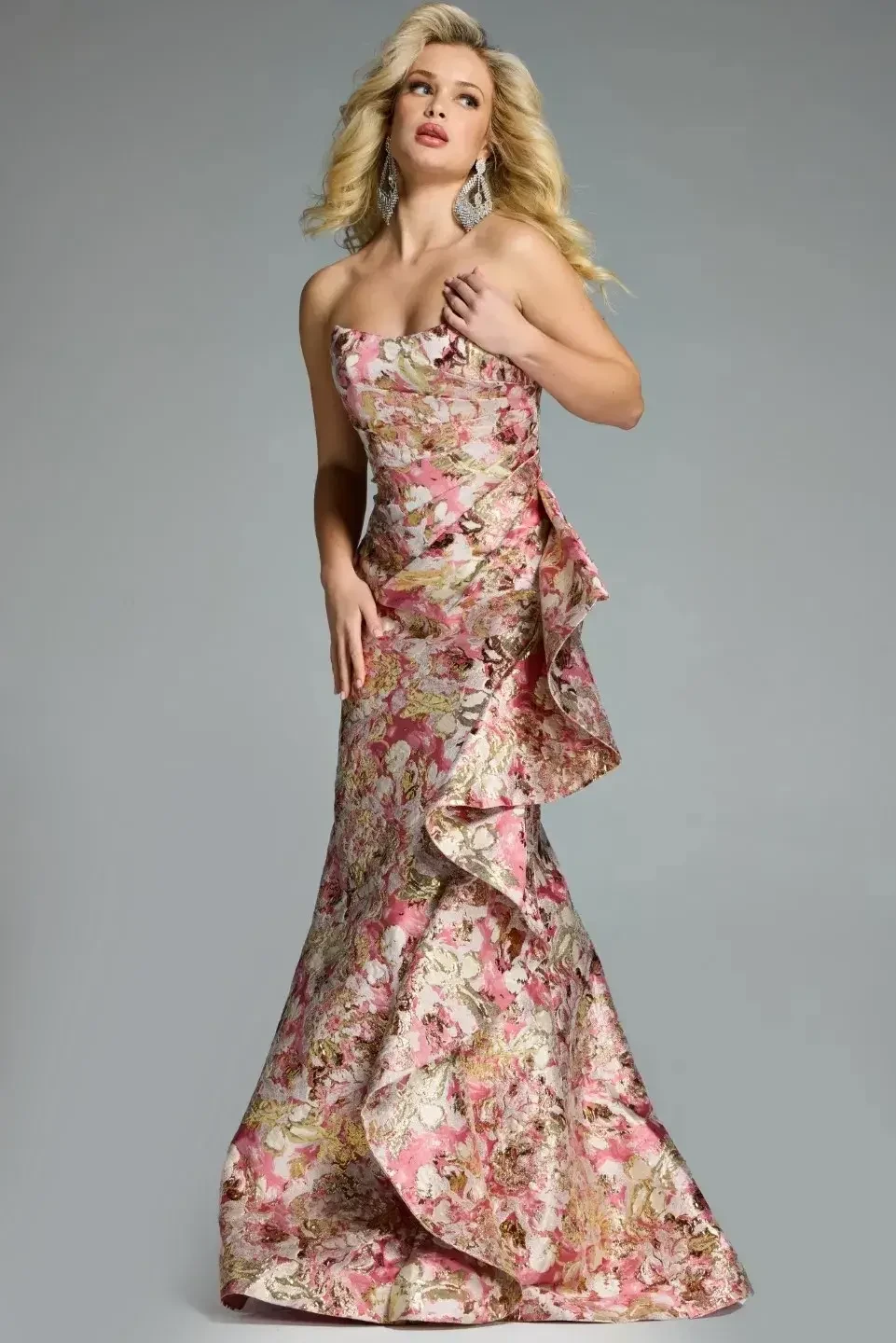 A woman wearing a floral strapless gown with a ruffled detail on the side, standing elegantly against a neutral background.