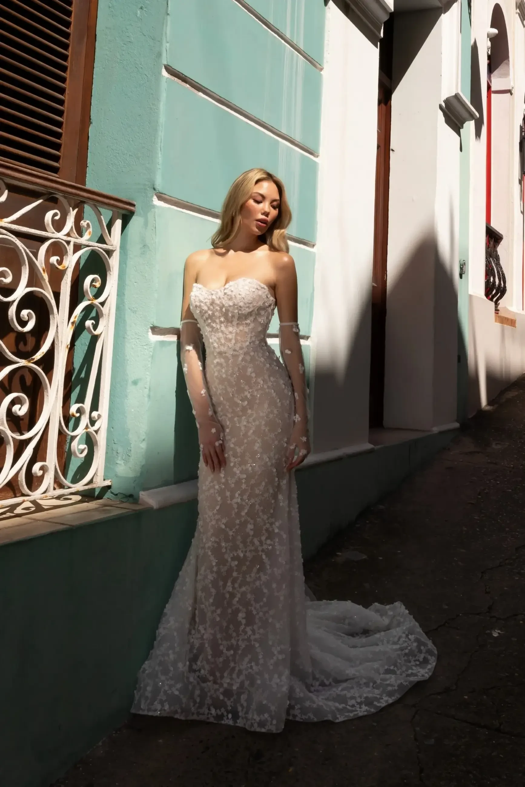 A woman in a white, intricately designed gown poses against a brightly colored wall, with sunlight illuminating her features.