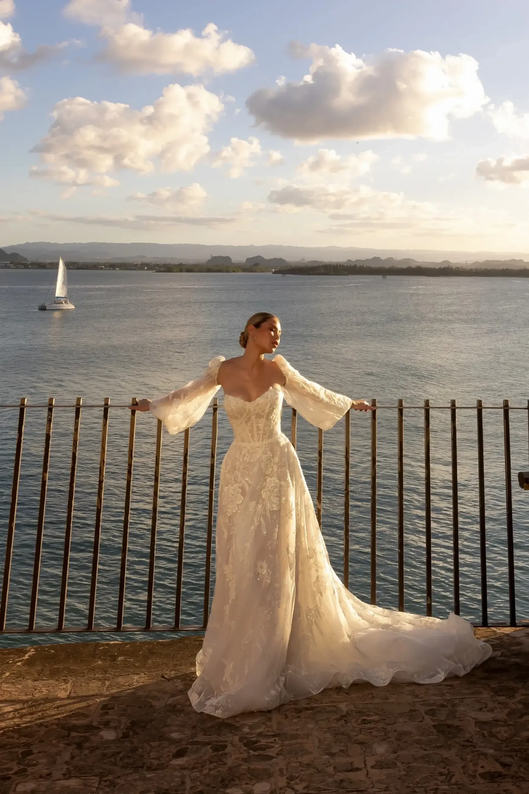 A woman in a flowing white gown stands by a railing with her arms outstretched, overlooking a scenic body of water under a cloudy sky. A sailboat is visible in the background.