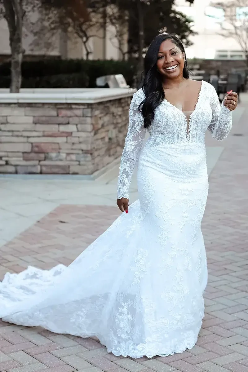 A woman wearing a long-sleeved, v-neck wedding dress with lace detailing, smiling while walking on a brick path.