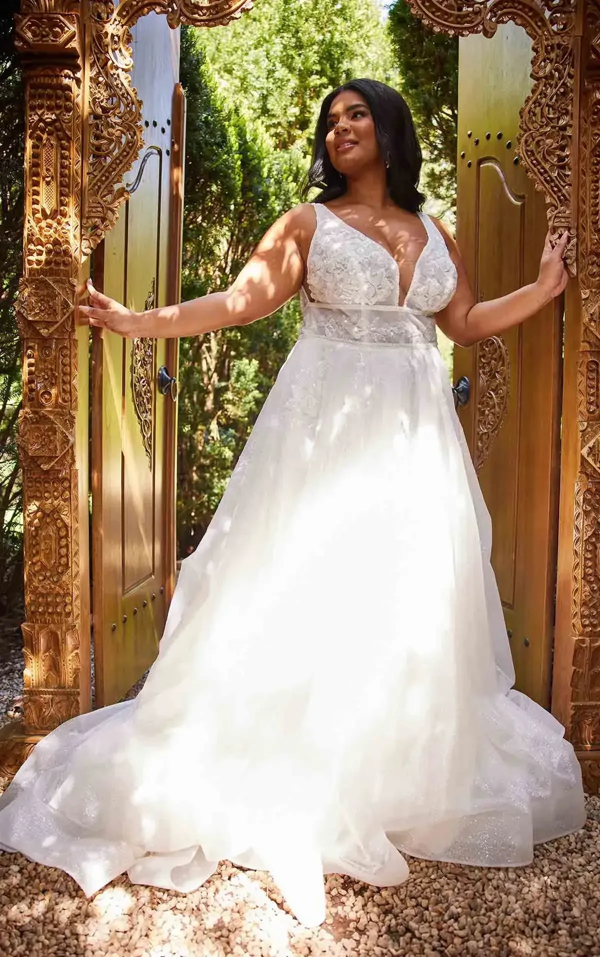 A woman is standing elegantly in a white wedding dress, framed by ornate wooden doors surrounded by greenery.