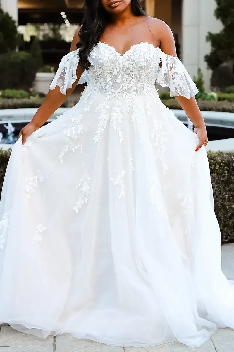 A person wearing a white wedding dress with off-the-shoulder sleeves and floral embellishments, standing outdoors with a fountain in the background.