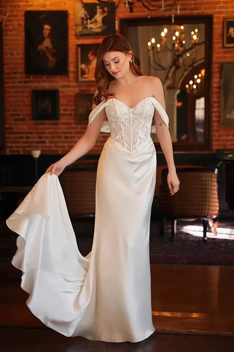 A woman in a white off-the-shoulder gown stands indoors, holding the train of her dress. The background features brick walls and framed artwork.