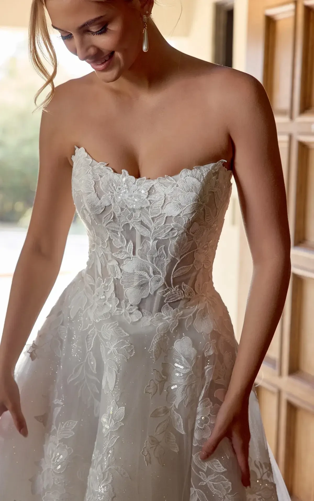 A woman in a strapless, intricately detailed wedding dress with floral motifs, smiling as she looks down at her outfit.