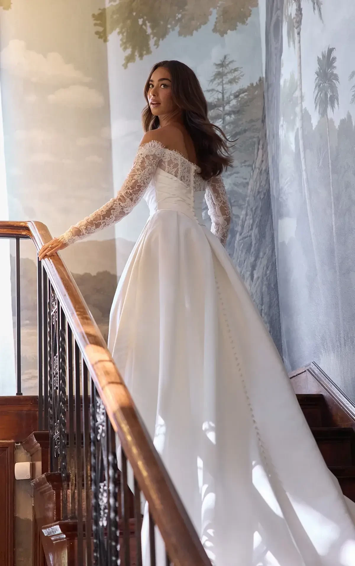 A woman in a white wedding dress with lace sleeves stands on a staircase, looking back over her shoulder. The background features a mural of trees and clouds.