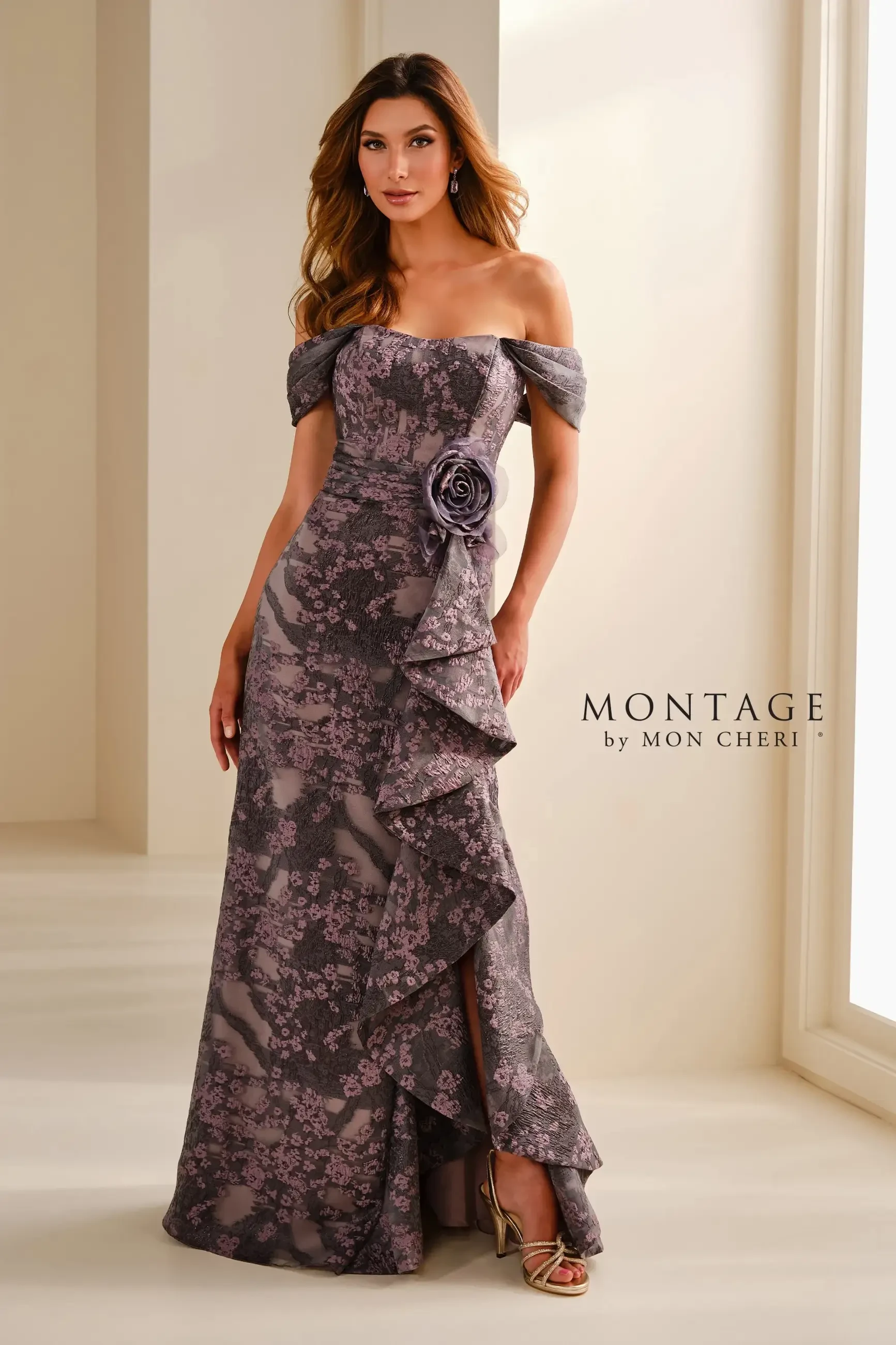 A woman wearing an elegant, off-the-shoulder gown with floral patterns and a ruffled design. The dress features a large flower detail at the waist and is paired with strappy heels. The setting appears to be well-lit and modern.